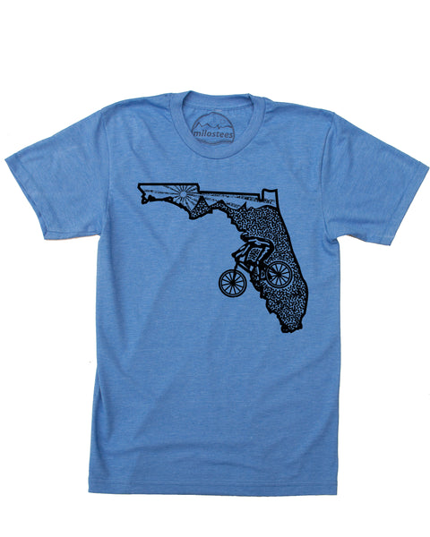This Bicycle Florida tee features a bold black graphic on a classic blue shirt. The design showcases a cyclist cruising across Florida's rolling hills with a stunning sunset stretching across the Panhandle. It's the perfect way to represent your love for the Sunshine State's cycling scene. Available in sizes Small through XXL.