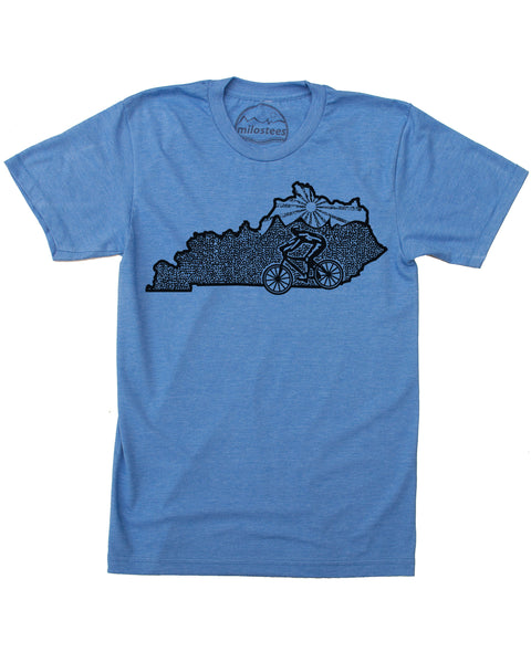 A heather blue T-shirt featuring a cyclist riding their bike across the state of Kentucky. The top third of the shirt depicts Kentucky's rolling hills with a setting sun in the background. Available in sizes small, medium, large, extra large, and double extra large.