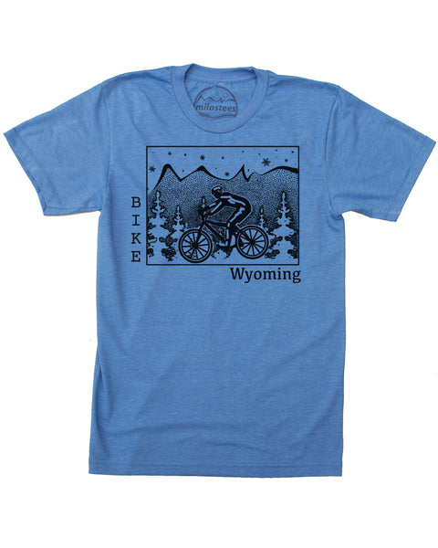 Black ink on a blue t-shirt depicts a cyclist riding across Wyoming. Pine trees surround the cyclist, snowflakes fall from the sky, and our mountain logo sits atop the state. "Bike" is written vertically along the left border of Wyoming, and "Wyoming" rests on the bottom right. 
