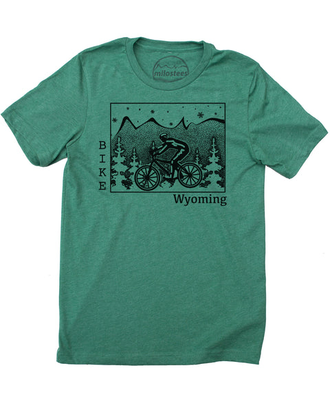 Black ink on a green t-shirt depicts a cyclist riding across Wyoming. Pine trees surround the cyclist, snowflakes fall from the sky, and our mountain logo sits atop the state. "Bike" is written vertically along the left border of Wyoming, and "Wyoming" rests on the bottom right. 