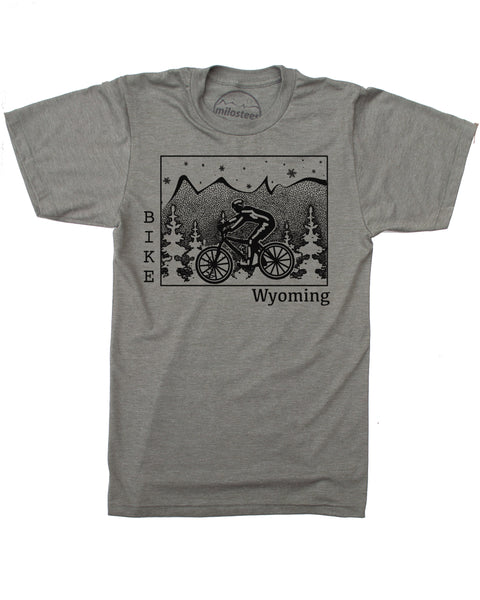 Black ink on a army green t-shirt depicts a cyclist riding across Wyoming. Pine trees surround the cyclist, snowflakes fall from the sky, and our mountain logo sits atop the state. "Bike" is written vertically along the left border of Wyoming, and "Wyoming" rests on the bottom right. 