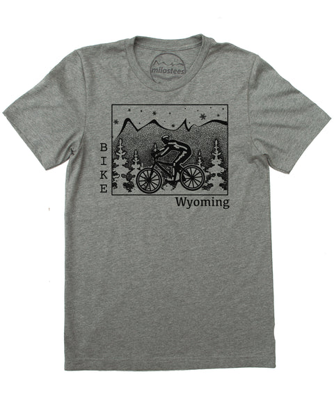 Black ink on a grey t-shirt depicts a cyclist riding across Wyoming. Pine trees surround the cyclist, snowflakes fall from the sky, and our mountain logo sits atop the state. "Bike" is written vertically along the left border of Wyoming, and "Wyoming" rests on the bottom right. 