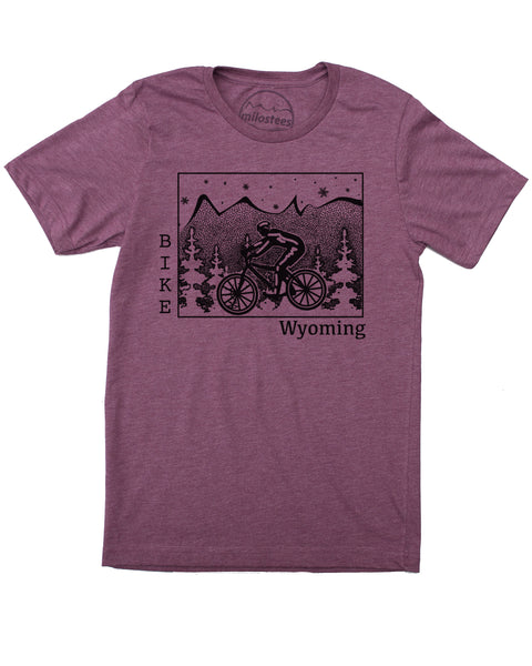 Black ink on a plum t-shirt depicts a cyclist riding across Wyoming. Pine trees surround the cyclist, snowflakes fall from the sky, and our mountain logo sits atop the state. "Bike" is written vertically along the left border of Wyoming, and "Wyoming" rests on the bottom right. 